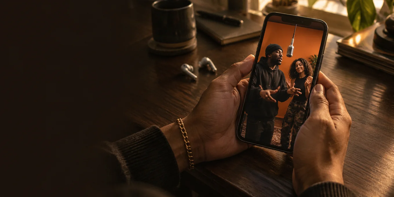 Hands hold a phone showing two performers in an orange studio.